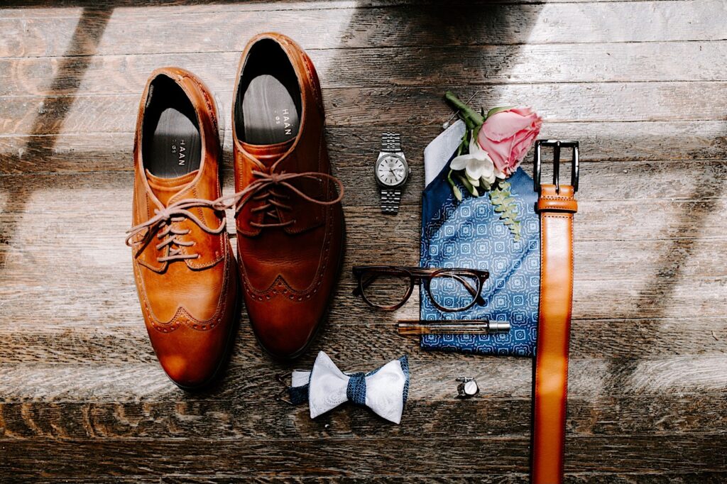 Detail photo of a wedding day flatlay featuring shoes, a bow tie, glasses, flowers, a belt, a watch, cufflinks, a handkerchief, and cologne