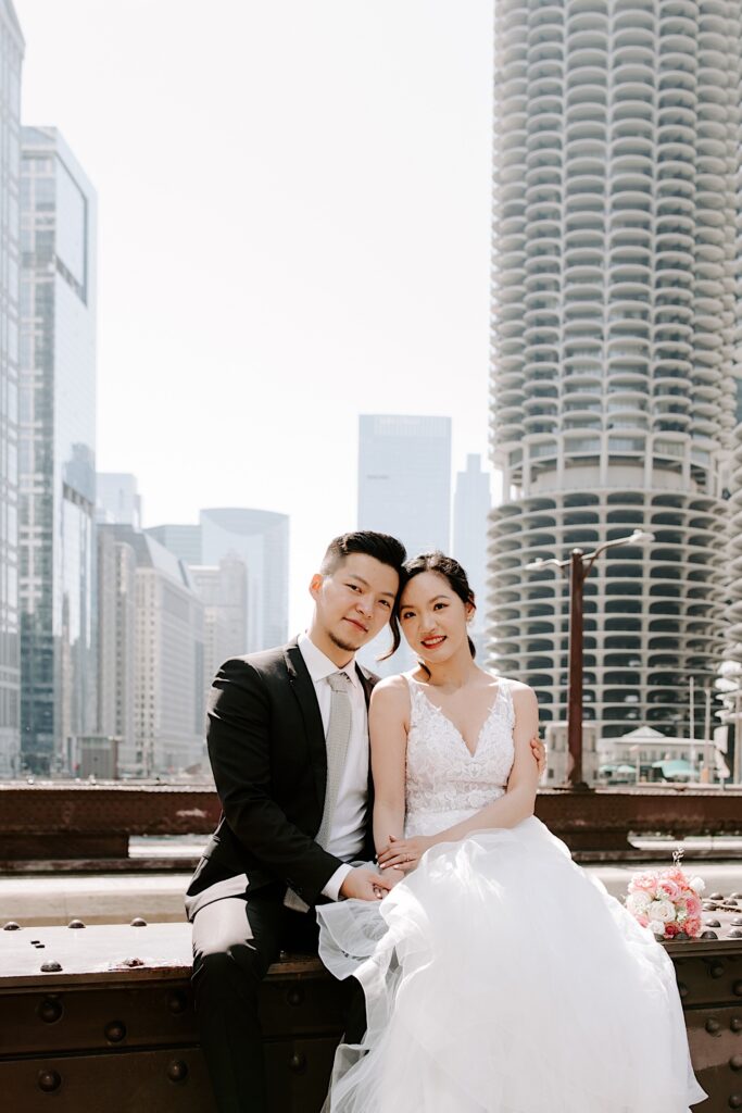 A bride and groom sit on a bridge in Chicago and embrace, behind them are buildings of the city