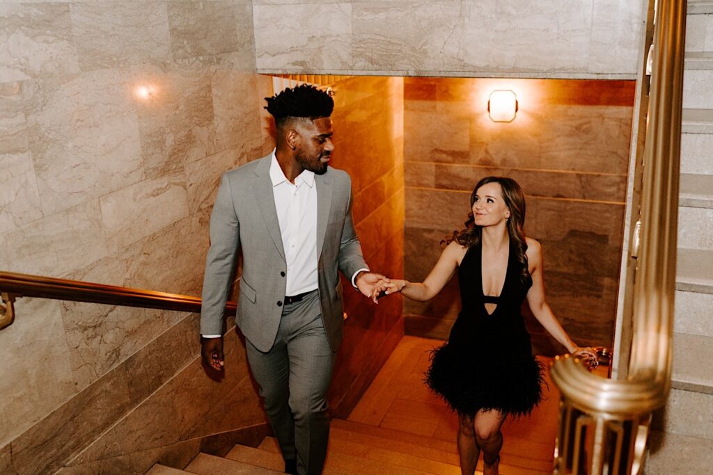 A couple indoors in a marble stairway walk hand in hand while smiling at one another during their engagement photos in Chicago