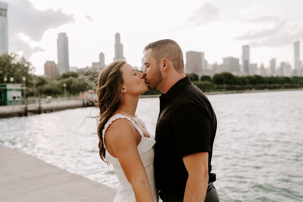 During their engagement session at Museum Campus a couple embrace and kiss one another in front of Lake Michigan and the Chicago skyline