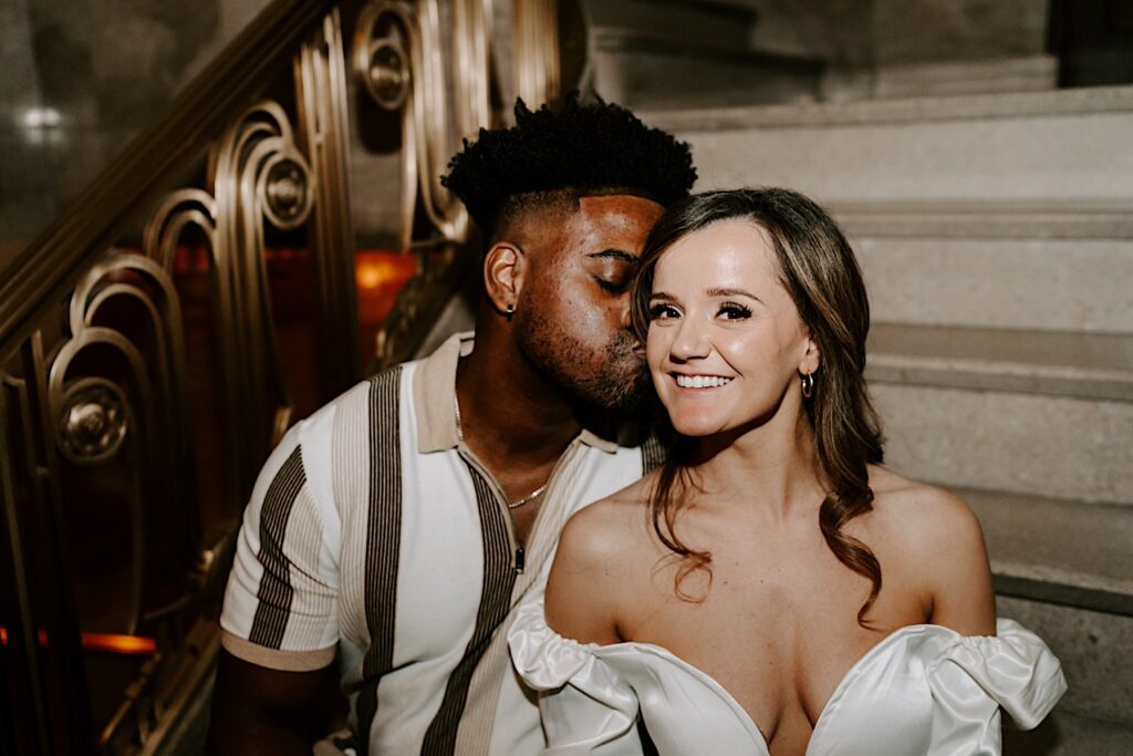 A couple sit on a marble staircase indoors in a building in Chicago taking engagement photos, the man is kissing the woman's cheek while she smiles at the camera