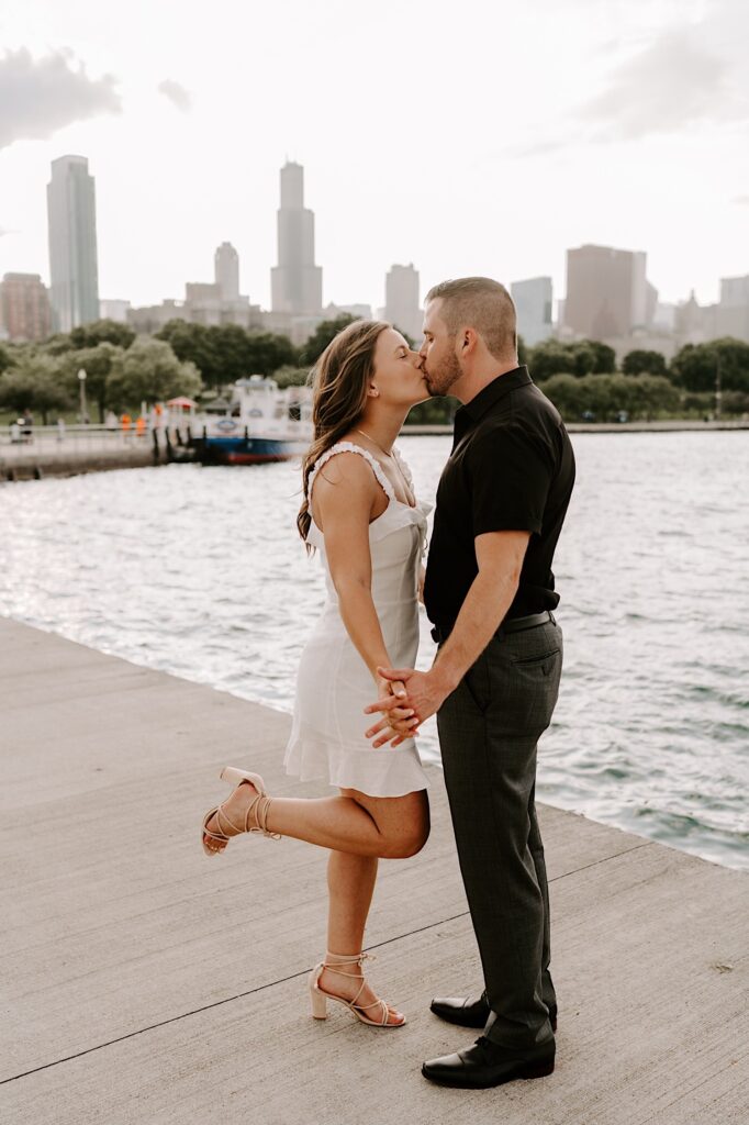 A couple kiss one another while holding hands in front of Lake Michigan and the Chicago skyline, the woman has one of her legs in the air