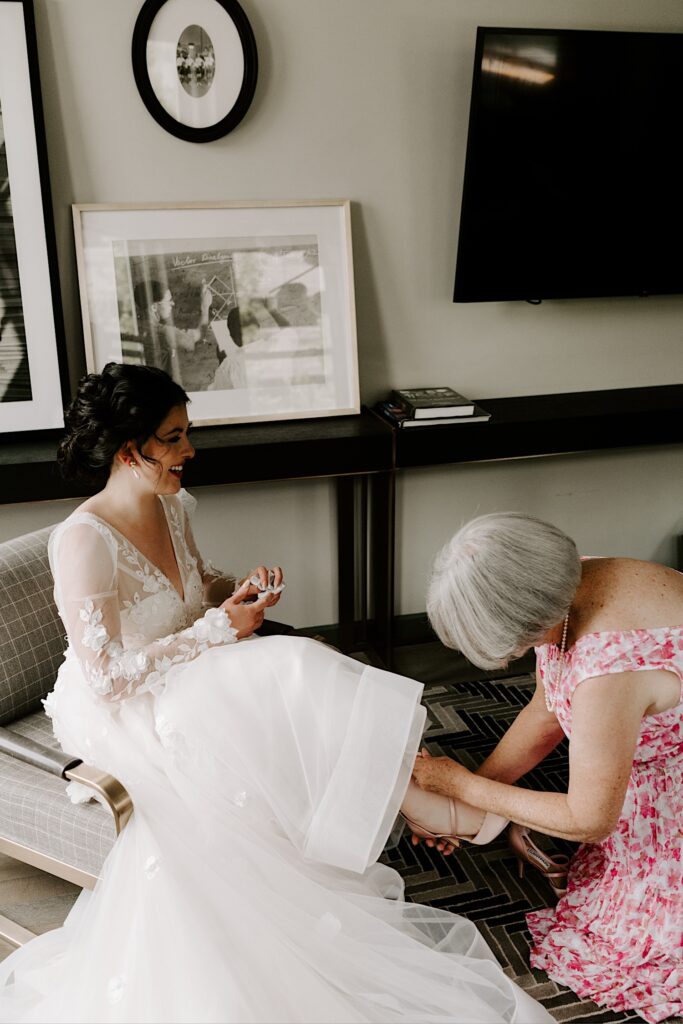 A bride sits and smiles as her mother helps put on her shoes before her wedding day