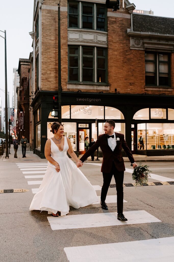 A bride and groom hold hands and smile at one another while crossing a street in Chicago