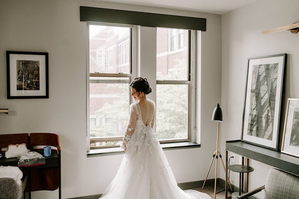 A bride in her wedding dress stands in front of a window and looks over her shoulder at the ground while smiling