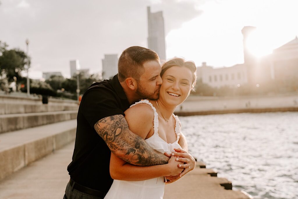 At their engagement session at Chicago's museum campus, a man hugs a woman from behind and kisses her on the cheek as she smiles at the camera while the sun sets behind them over Lake Michigan