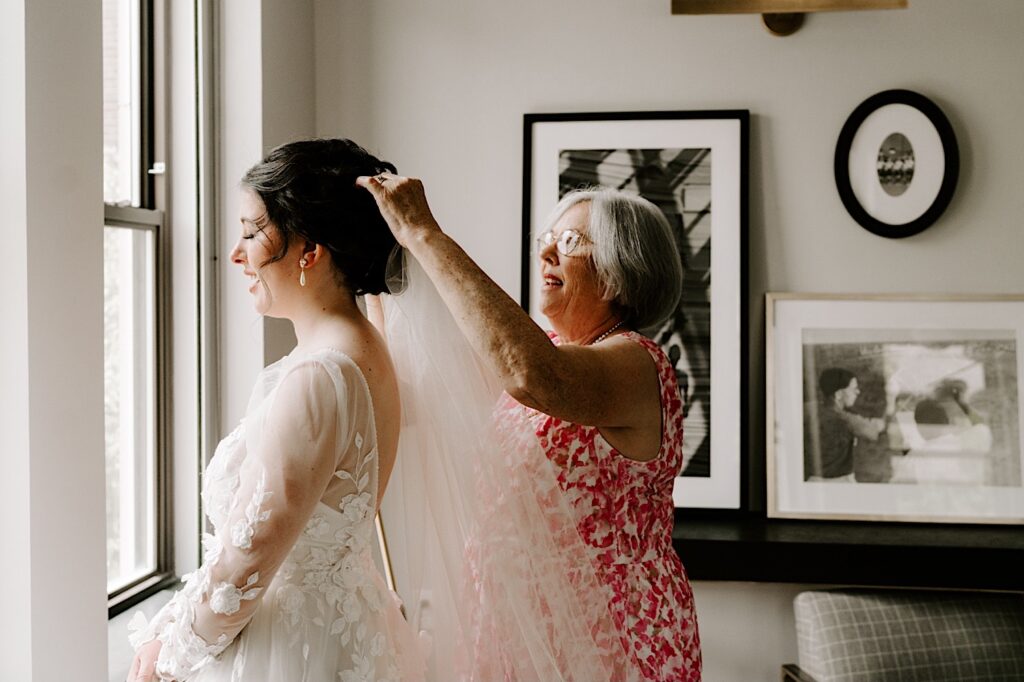 A bride in her wedding dress stands in a window and smiles as her mother stands behind her and adjusts her wedding veil