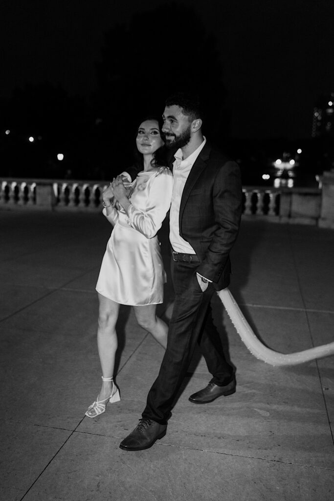 Black and white photo of a brie and groom walking outside at night on a large balcony, the bride smiles at the groom as he looks straight ahead