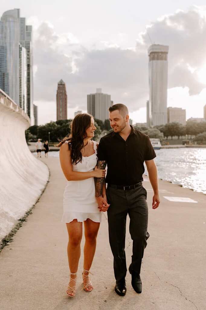 A couple hold hands and walk along the shore of Lake Michigan at Chicago's museum campus with the skyline behind them