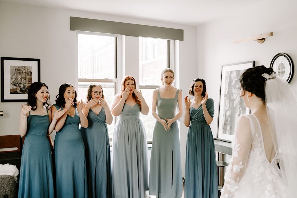 A bride with her back to the camera stands in her wedding dress in front of her six bridesmaids who all react seeing her in her wedding dress for the first time