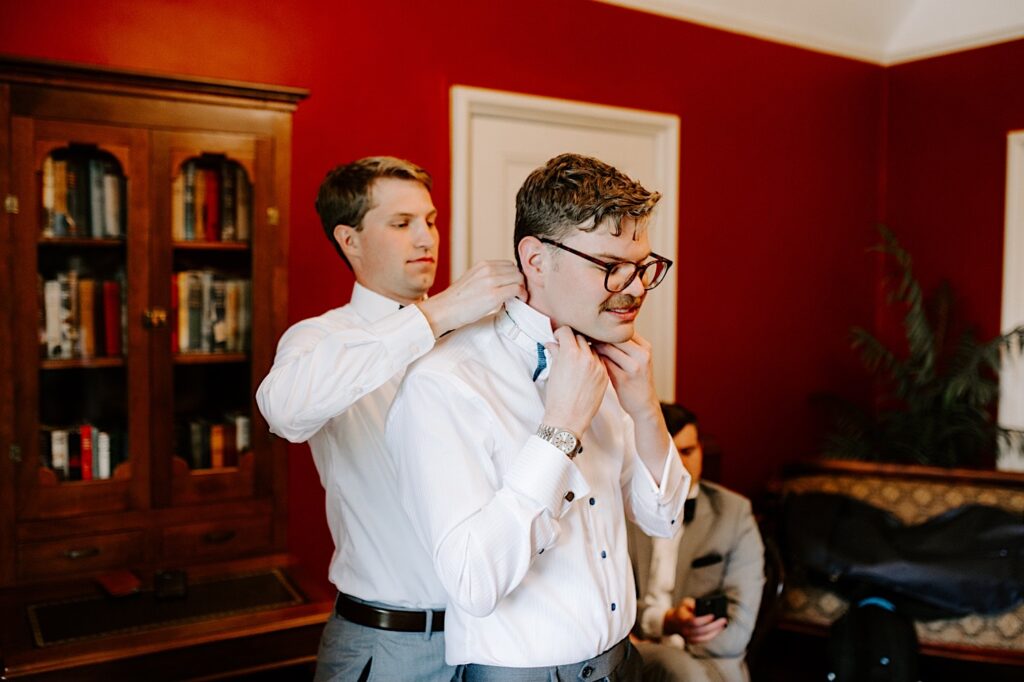 A groom adjusts his bow tie as one of his groomsmen helps adjust his collar behind him
