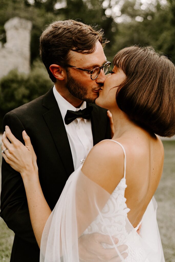 A bride and groom kiss and embrace outdoors with grass and trees behind them