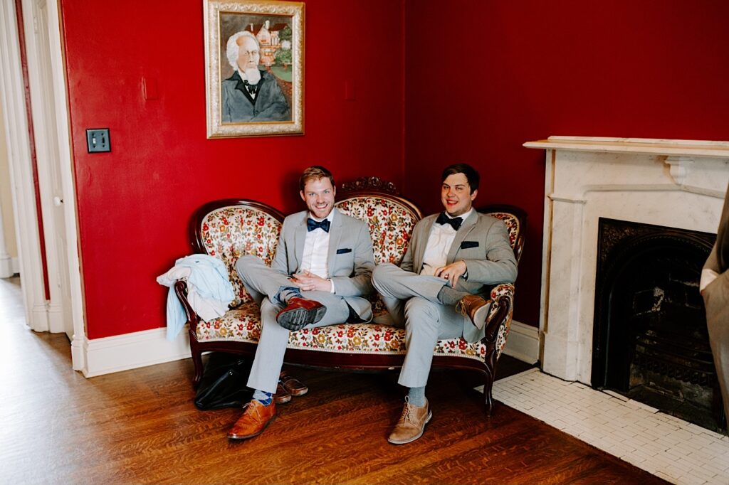 Two groomsmen sit on a floral couch in a red room in their wedding suits while smiling at the camera
