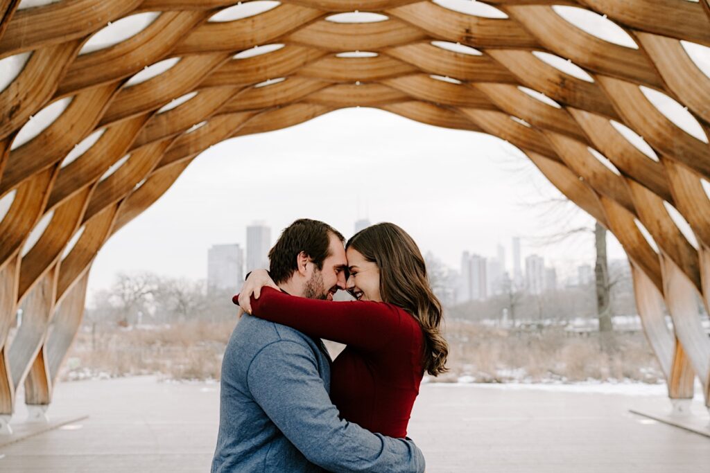 A couple embrace and smile at one another while standing underneath the "honeycomb" at Lincoln Park in Chicago during the winter as there is snow on the ground behind them
