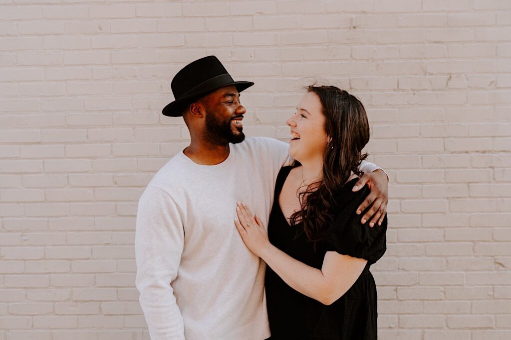 A couple smile at one another and embrace in front of a brick wall during their engagement photos, the woman has her hand on the man's chest showing off her engagement ring