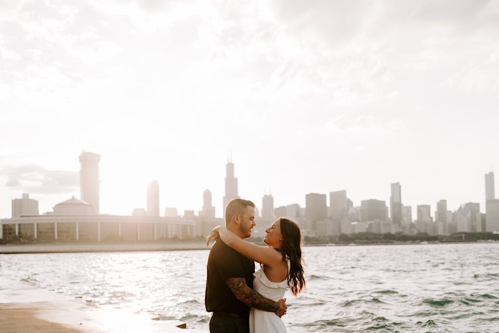 A couple embrace and smile at one another during their engagement session at Chicago's museum campus, behind them the sun sets over the Chicago skyline and Lake Michigan