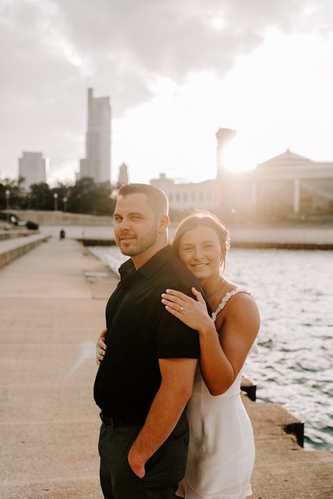 A woman hugs a man from behind as they both smile at the camera while the sun sets behind them over Lake Michigan and the Chicago skyline
