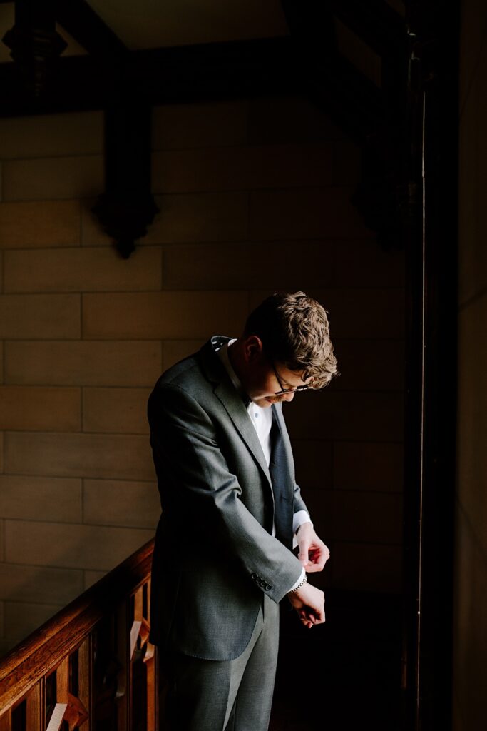 A groom standing in a dark room in front of a window adjusts his suit coat cuff while looking down