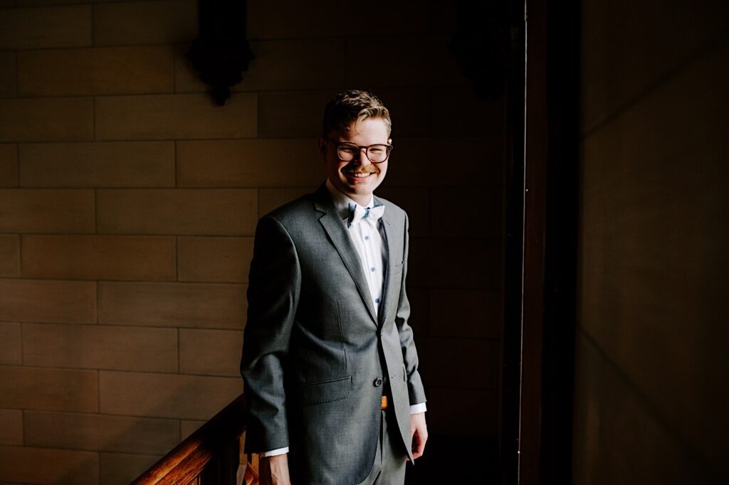A groom in his wedding suit stands in a dark room in front of a brick wall and smiles at the camera