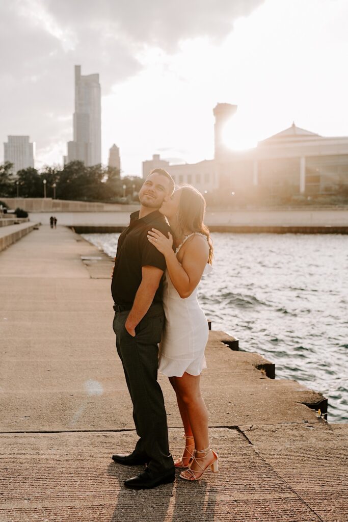 A woman hugs a man from behind and kisses his cheek as he smiles at the camera while the sun sets behind them over Lake Michigan and the Chicago skyline