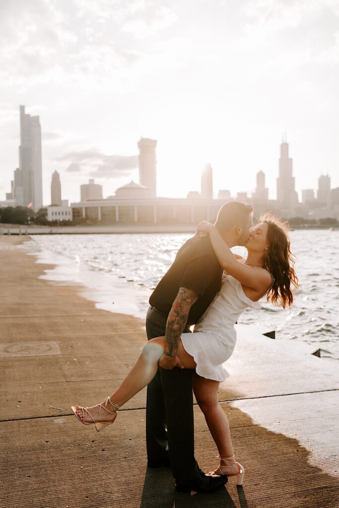 A man kisses a woman while holding her leg as she wraps her arms around him while the sun sets behind them over the Chicago skyline and Lake Michigan