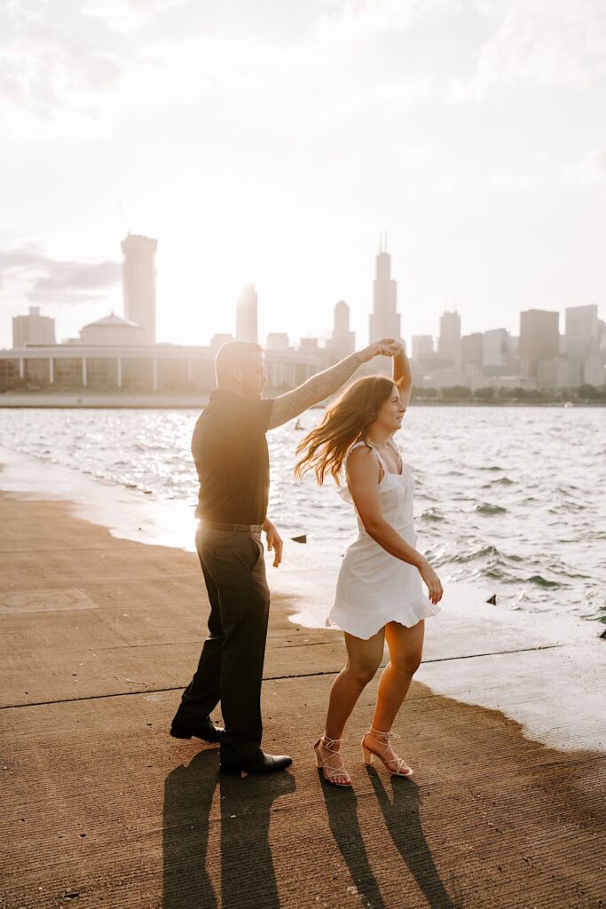 A woman spins while dancing with a man on the shore of Lake Michigan as the sun sets behind them over the Chicago skyline