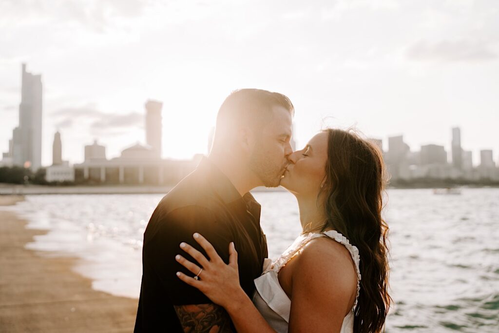 During their engagement session at Chicago's Museum Campus, a couple kiss one another and embrace while the sun sets behind them over Lake Michigan and the Chicago skyline