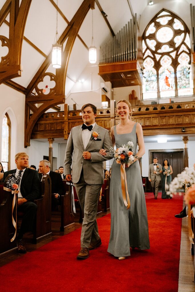 A groomsman and bridesmaid walk down the aisle of a church and smile before a wedding ceremony