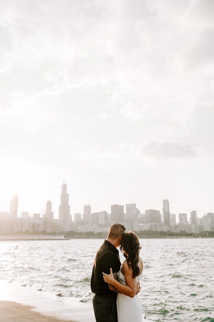 A couple embrace one another as they look away from the camera at the Chicago skyline and Lake Michigan