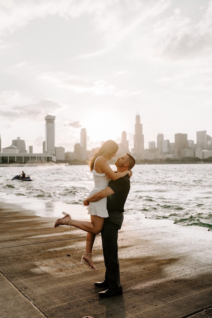 A man lifts a woman in the air on the shore of Lake Michigan with the sun setting behind them on the Chicago skyline