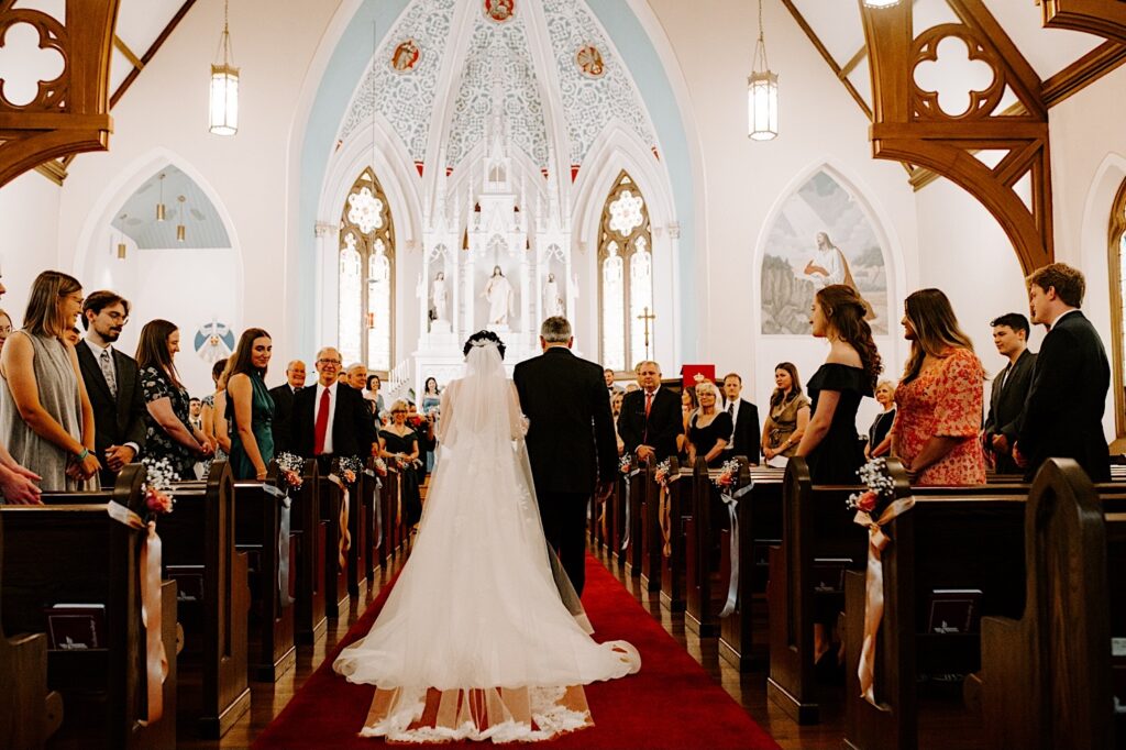 Inside a church a bride is walked down the aisle by her father away from the camera during her wedding ceremony