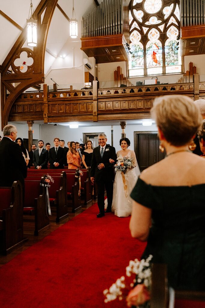 A bride is walked down the aisle of a church by a father as guests stand around them during her wedding ceremony