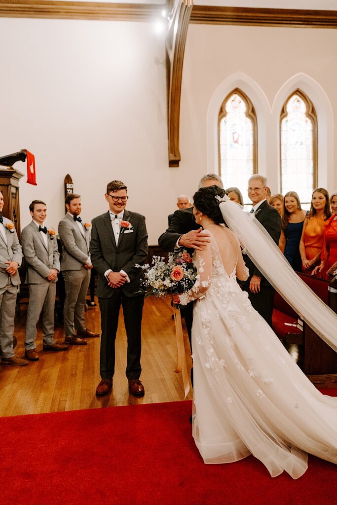 A groom smiles as the bride is hugged by her father after he walked her down the aisle during their church ceremony