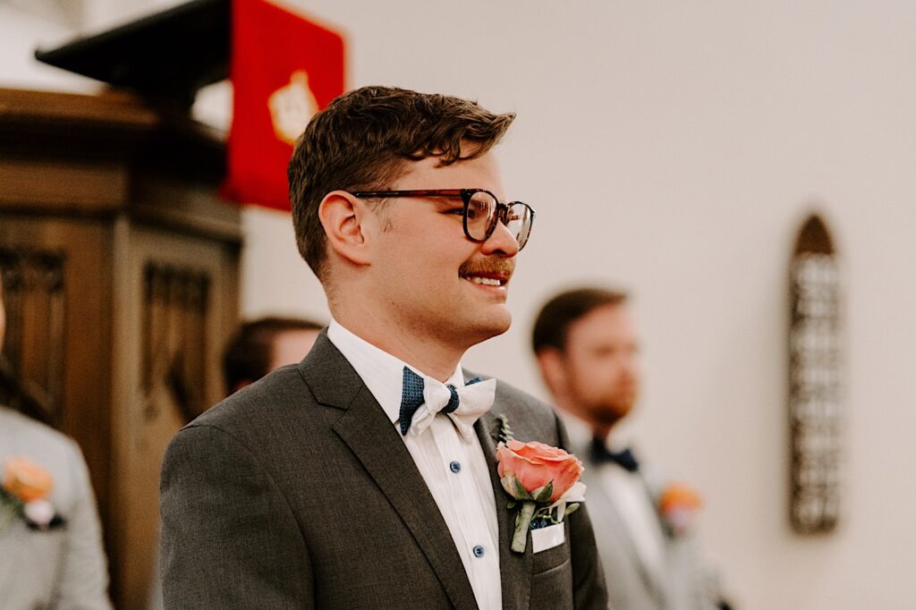 A groom smiles as he looks down the aisle towards his bride during their wedding ceremony