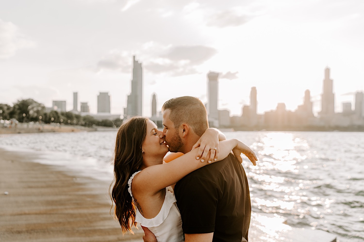 A couple embrace and kiss one another during their engagement session at Chicago's Museum Campus as the sun sets behind them over the Chicago skyline and Lake Michigan