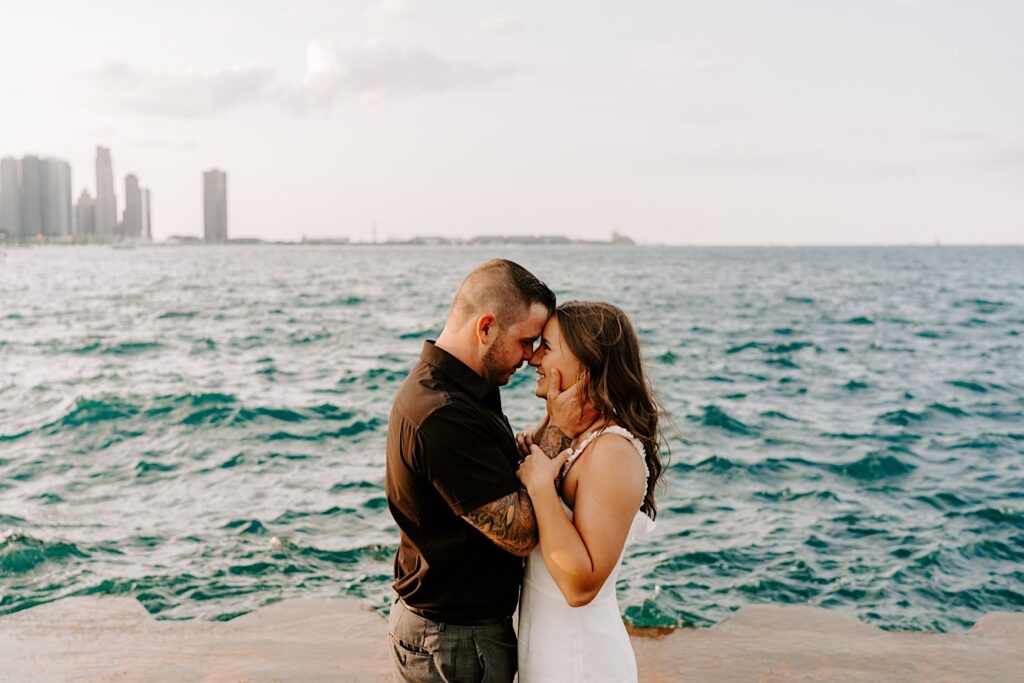 During their engagement session at Chicago's Museum Campus, a couple embrace and smile at one another while touching their noses together in front of Lake Michigan and the Chicago skyline
