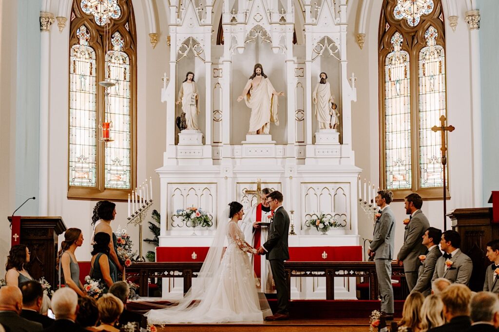 During a ceremony in a church a bride and groom stand together and hold hands as the pastor speaks