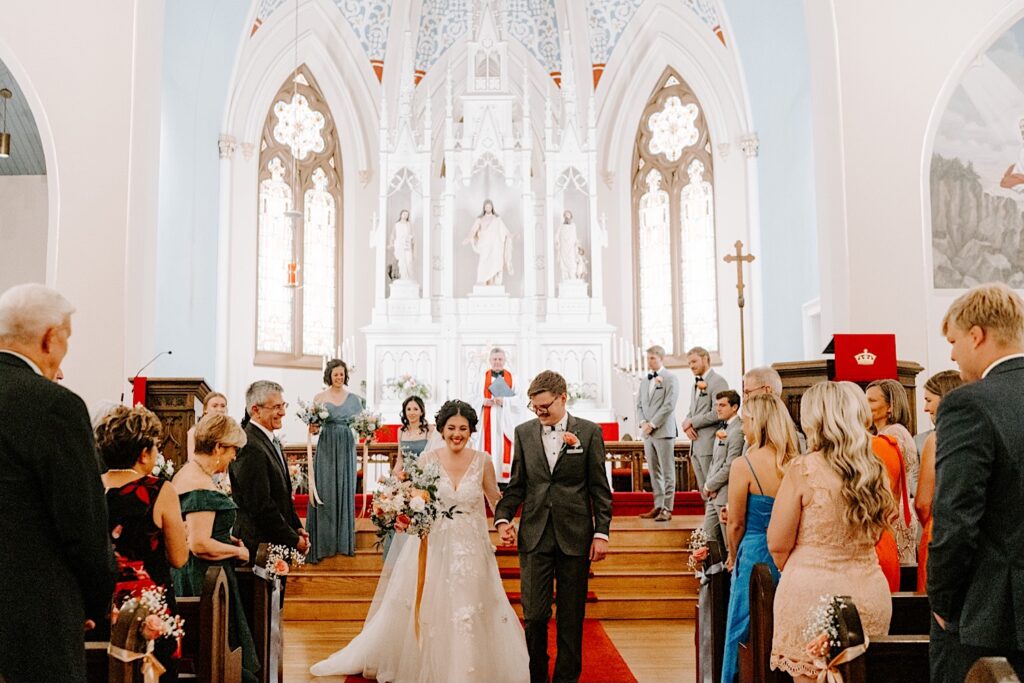 A bride and groom walk hand in hand and smile while walking down the aisle of a church together after their wedding ceremony