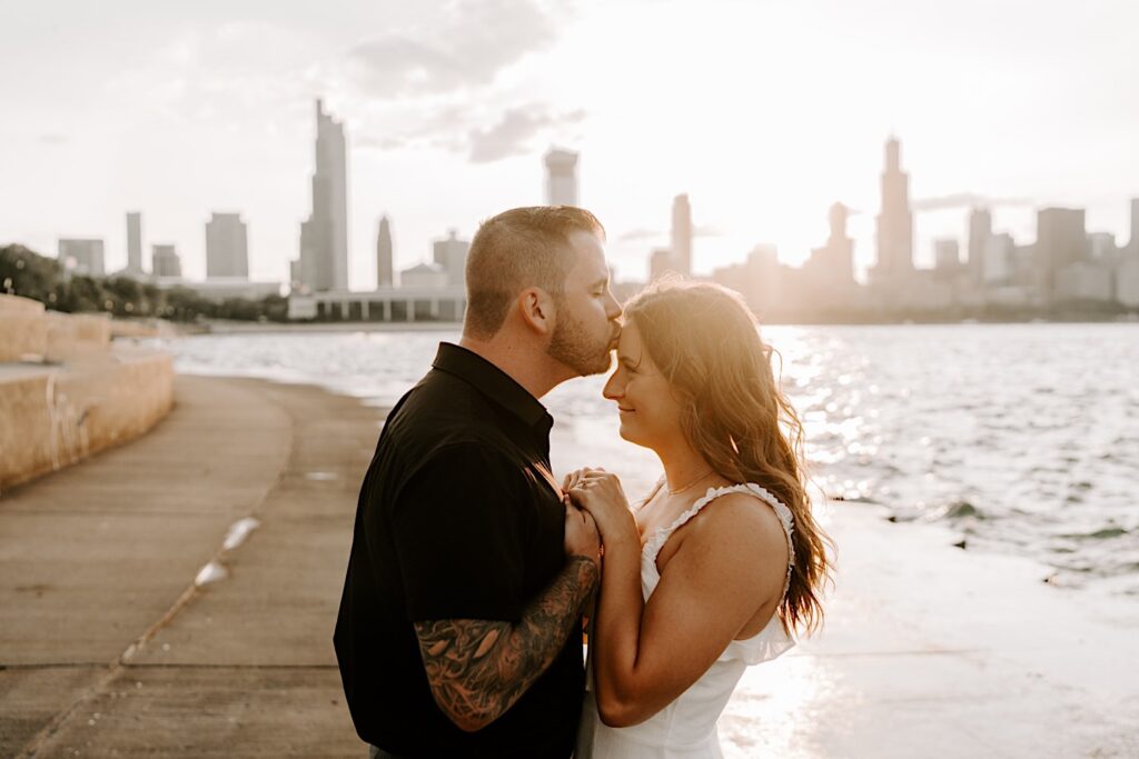 During their engagement session at Chicago's Museum Campus, a man kisses a woman on her forehead while she smiles at him in front of Lake Michigan and the Chicago skyline at sunset