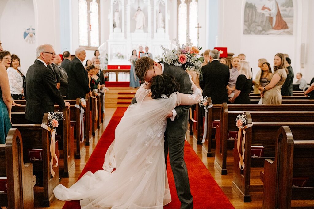 A groom dips and kisses a bride while they walk down the aisle of a church after their wedding ceremony