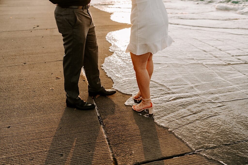 Waist down photo of a man and a woman's legs with water from Lake Michigan splashing up over the concrete barrier and underneath their feet