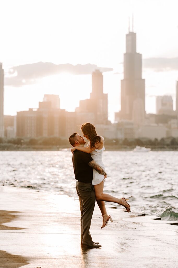 A man lifts a woman in the air as they look at one another in front of the Chicago skyline and Lake Michigan during sunset