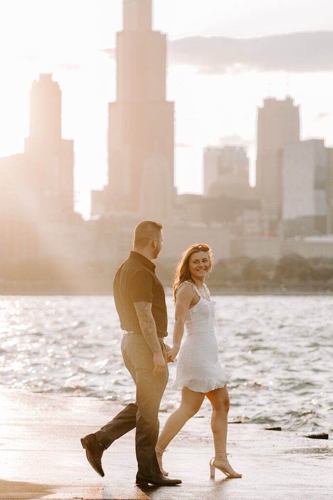 A couple walk hand in hand and look at one another in front of the Chicago skyline and Lake Michigan during sunset