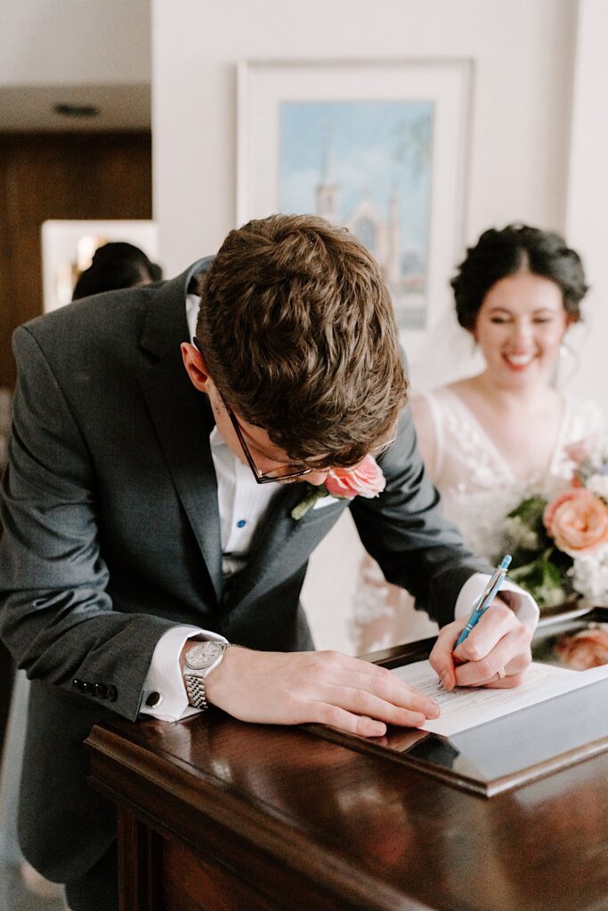A groom signs his wedding papers as the bride smiles standing behind him
