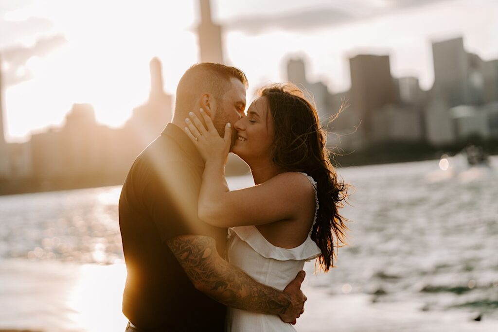 During their engagement session at Chicago's Museum Campus, a couple embrace and the woman smiles as the man kisses her cheek, the sun is setting on the Chicago skyline and Lake Michigan behind them