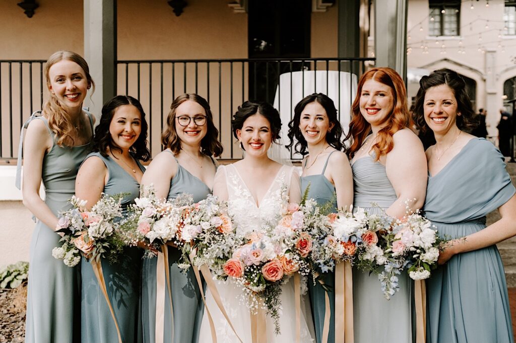 A bride and her six bridesmaids stand together holding bouquets and all smile at the camera outside the wedding venue the Fowler House Mansion