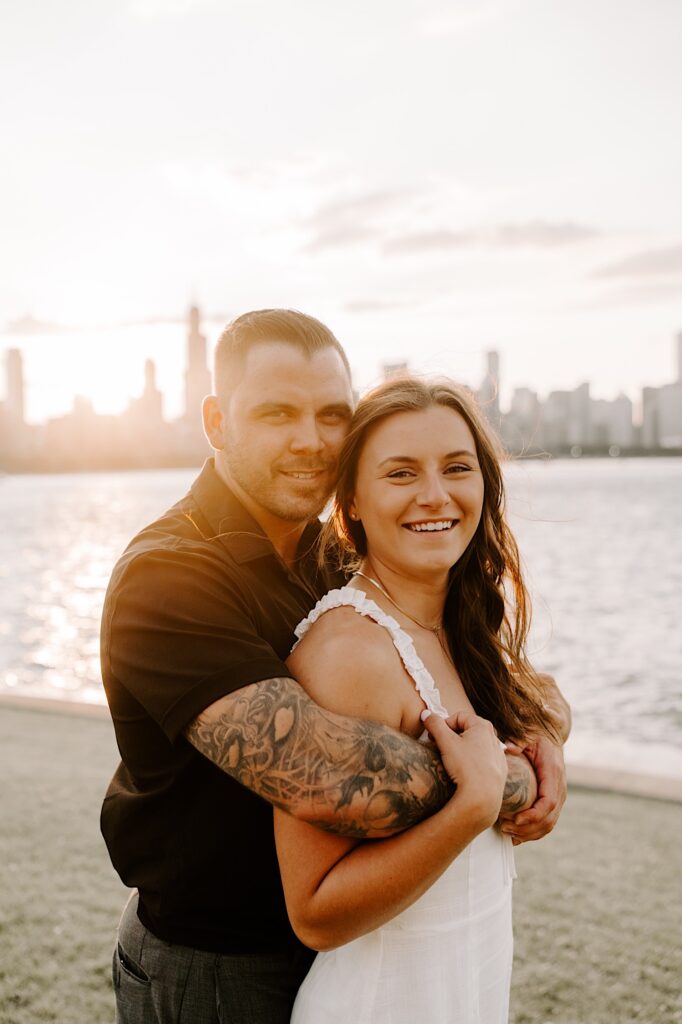 A man hugs a woman from behind and they both smile at the camera while standing in front of Lake Michigan and the Chicago skyline during sunset