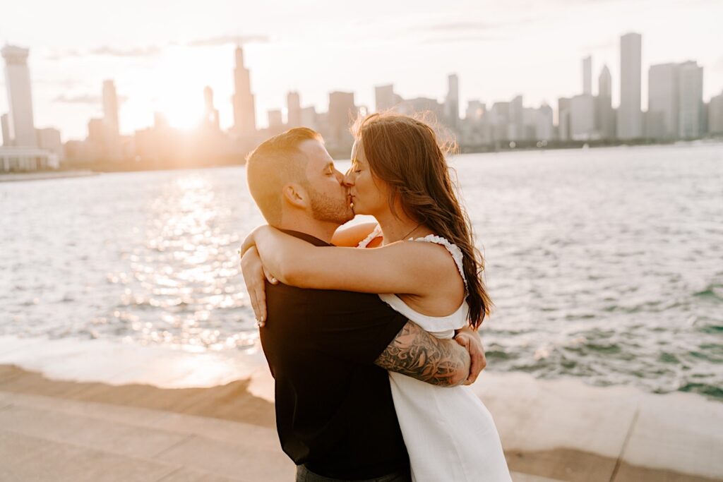 During their engagement session at Chicago's Museum Campus, a couple embrace and kiss one another during sunset with the Chicago skyline and Lake Michigan behind them
