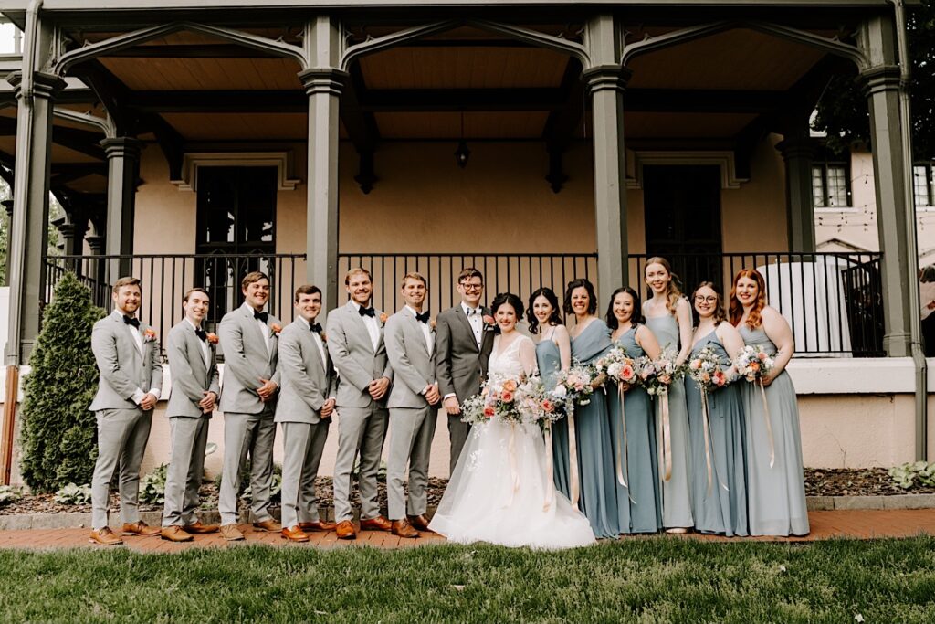 A bride and groom stand together and smile at the camera with either of their wedding parties on either side of them smiling at the camera as well outside their wedding venue the Fowler House Mansion