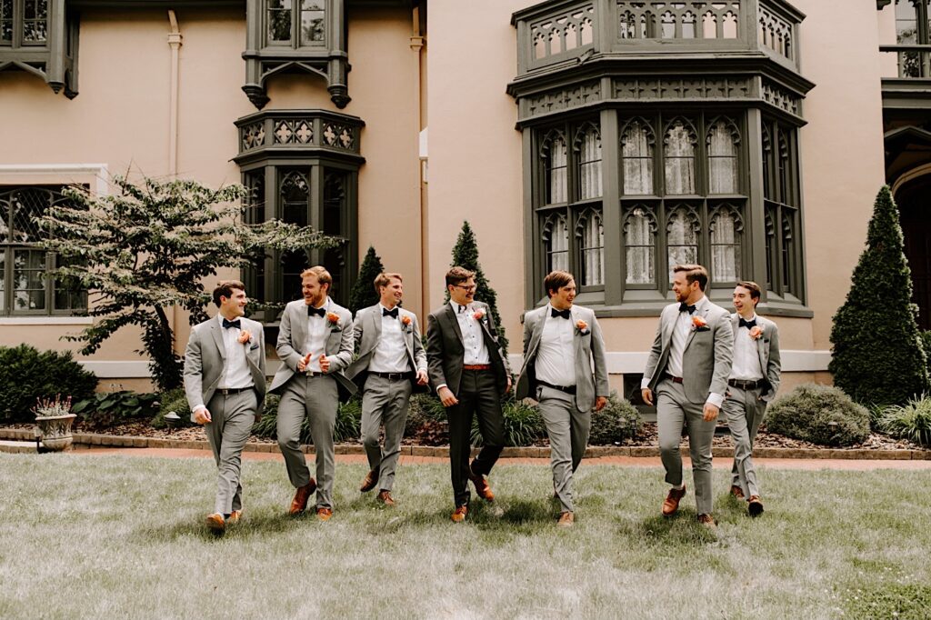 Outside the Fowler House Mansion a groom and his groomsmen walk towards the camera and smile while laughing at one another
